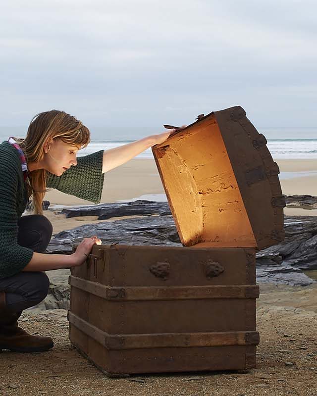 A woman looks inside a treasure chest with gold on a deserted beach.